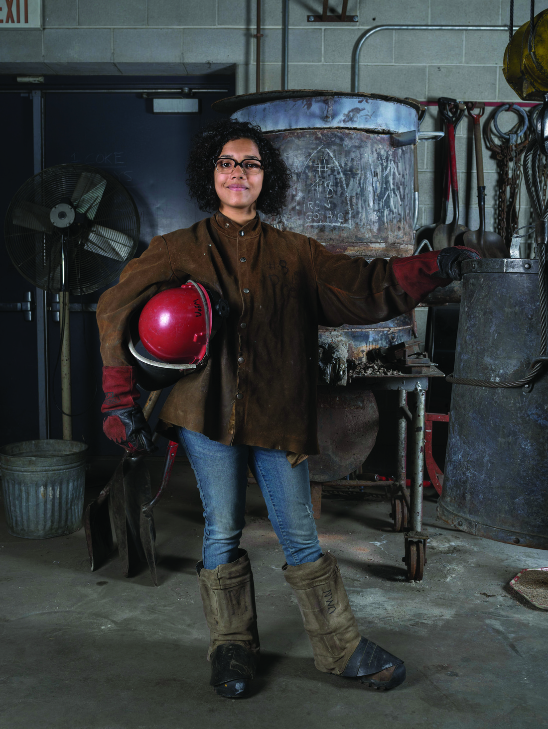 Woman in foundary wearing heavy boots and jacket, carrying a welding mask